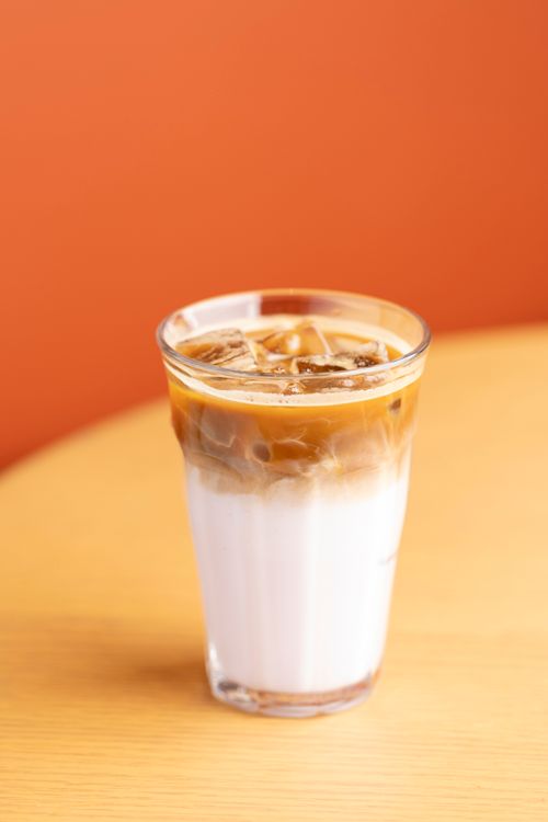 Free Iced latte glass on table with vibrant orange backdrop, perfect for summer vibes. Stock Photo