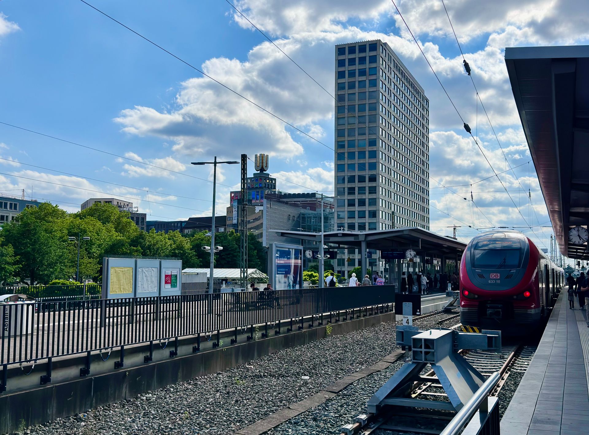 A bustling urban train station with a red train and city skyscrapers on a sunny day.
