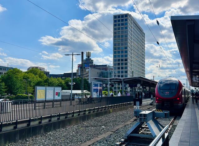 A bustling urban train station with a red train and city skyscrapers on a sunny day.