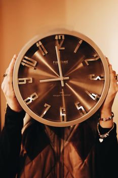 Person hides face behind a large brown wall clock in warm lighting.