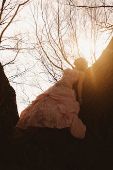 A bride in a pink gown leans against a tree with sunlight filtering through winter branches.