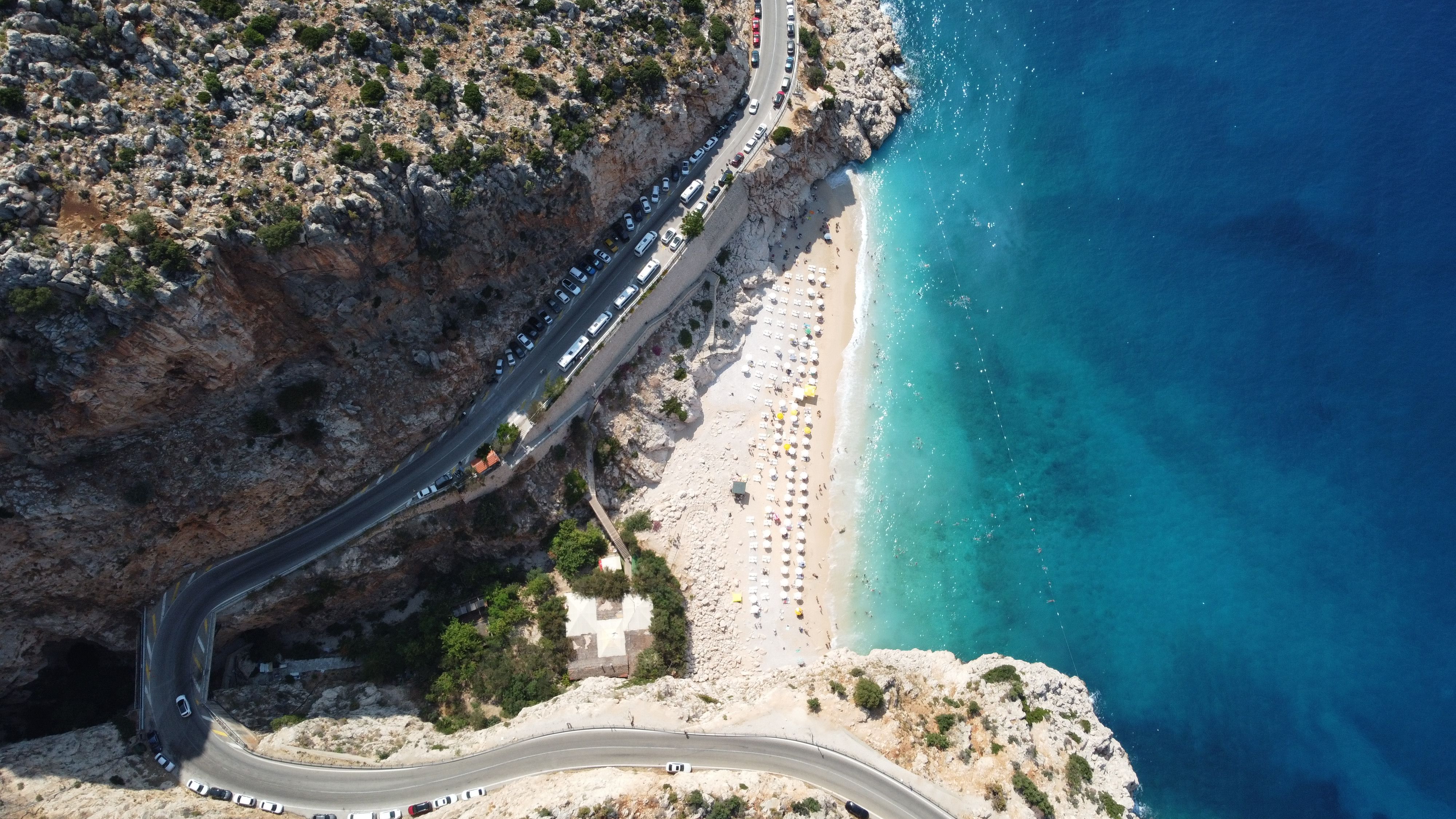 Breathtaking aerial view of a coastal beach with clear blue waters and a winding road along rocky cliffs. - Photo by Ahmet KADIOĞLU on Pexels