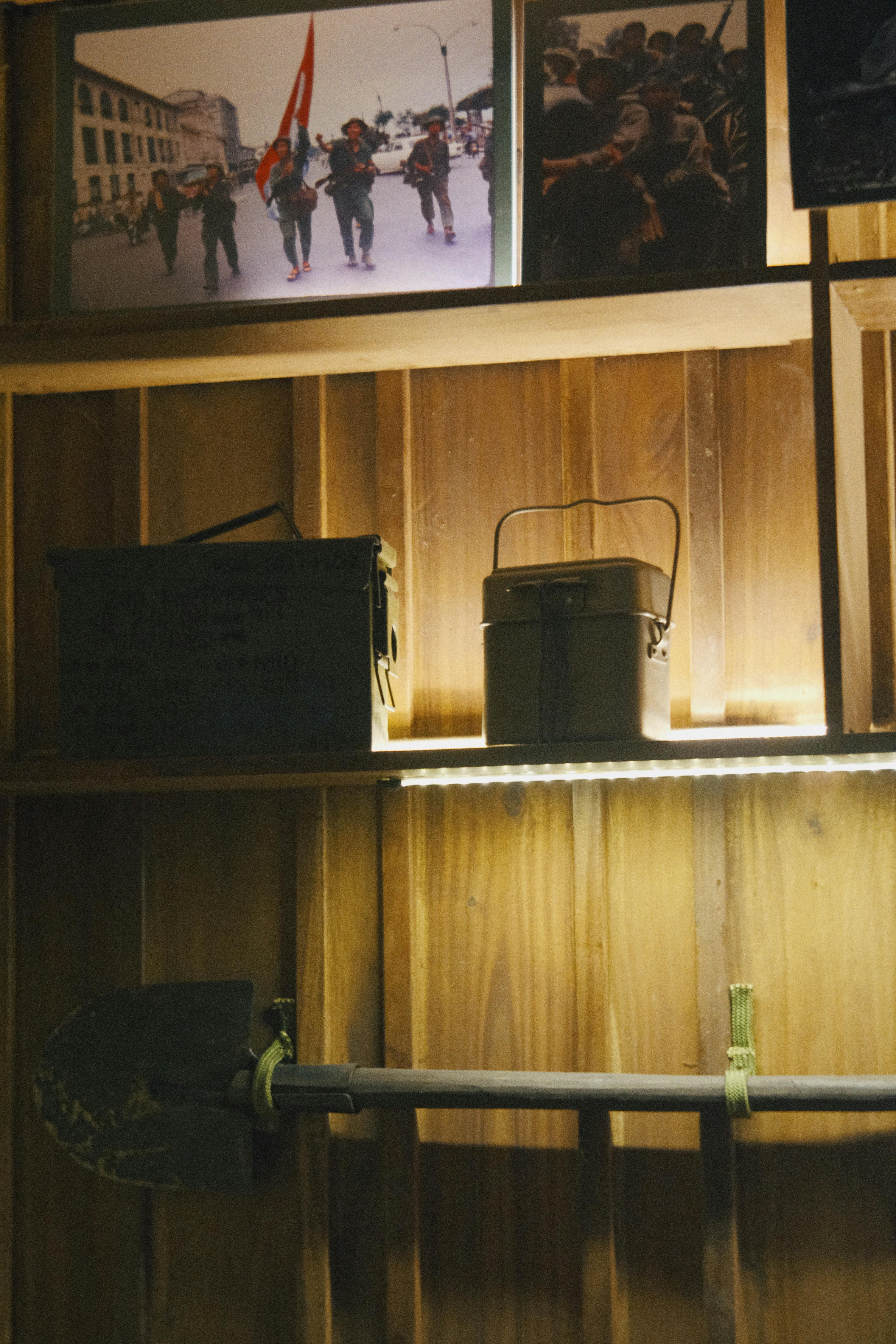 Wooden shelf with vintage military items, including a shovel and ammo boxes, under framed photographs.