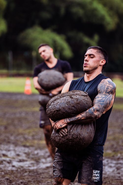 Free Two men vigorously carry weighted bags in a muddy outdoor fitness challenge. Stock Photo