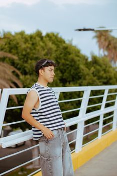 A young man in casual attire leans against a bridge railing, set in a lush outdoor environment.