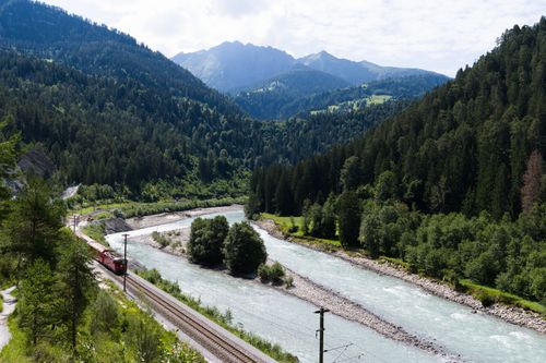 Free A picturesque view of a red train moving through a lush Swiss valley with a flowing river and mountains. Stock Photo