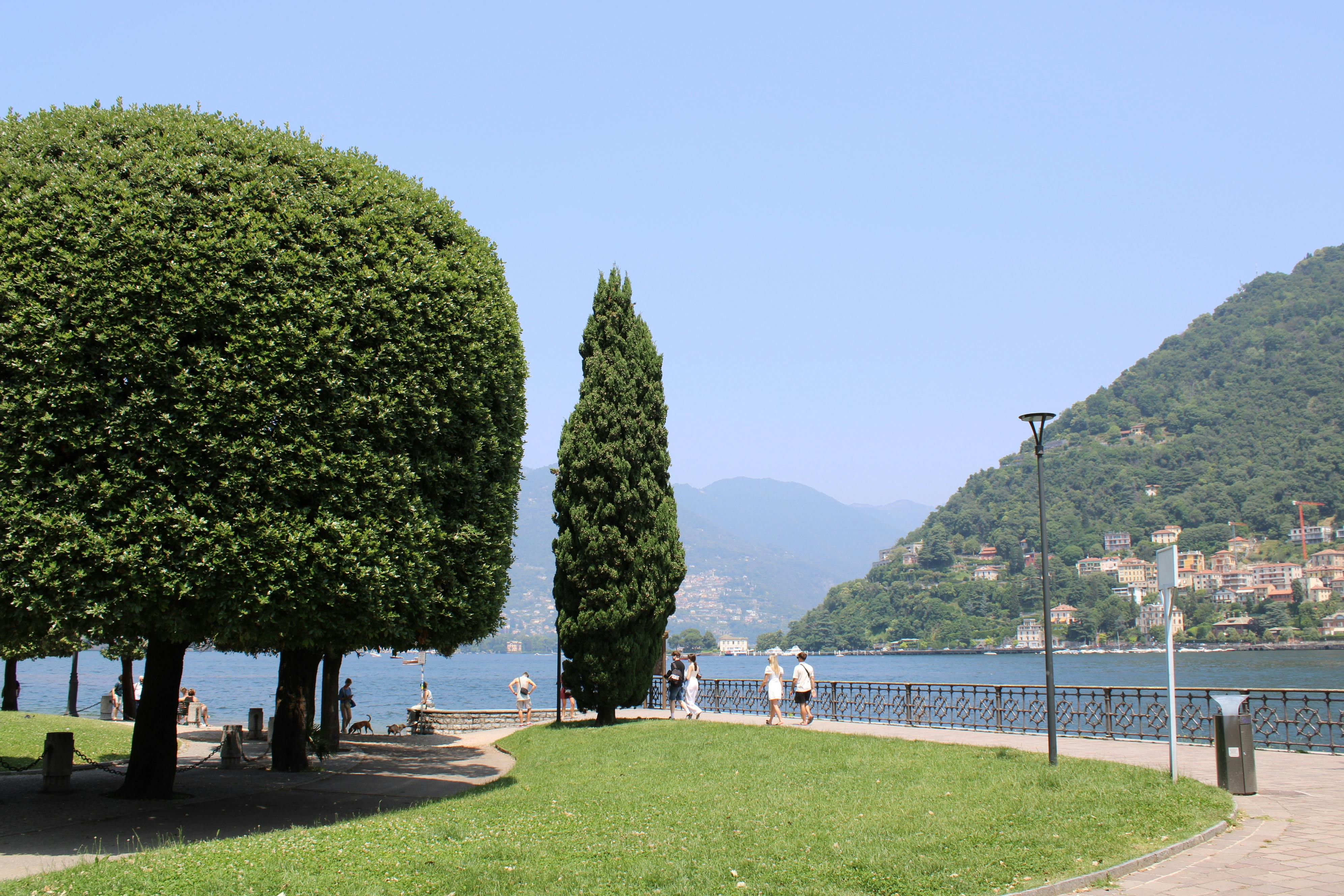 Scenic lakeside walking path along Lake Como with lush trees, mountain views, and calm waters — a peaceful promenade connecting the city of Como to Villa Olmo along the picturesque shoreline
