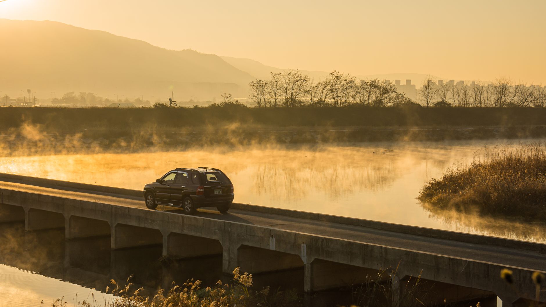 Car travels across a bridge over a misty river at sunrise, surrounded by serene landscapes.