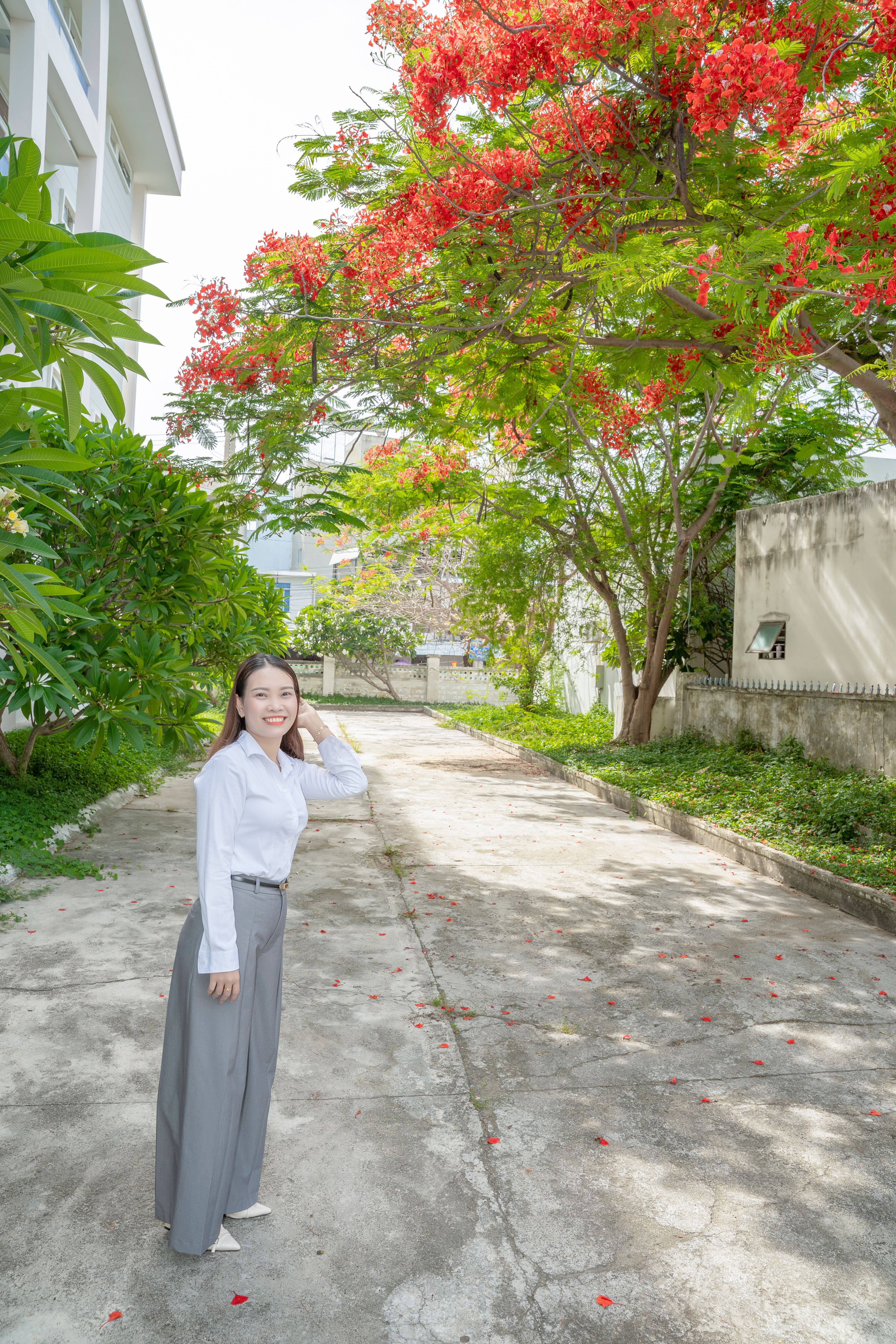 Adult woman smiles under a vibrant tree with red blooms, standing in a sunlit pathway outdoors.