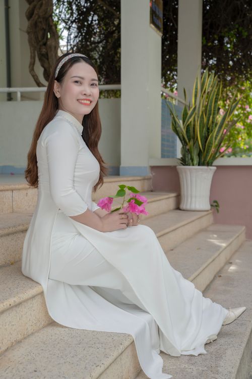 Free Woman in traditional white Ao Dai sitting on steps holding pink flowers, embracing elegance. Stock Photo
