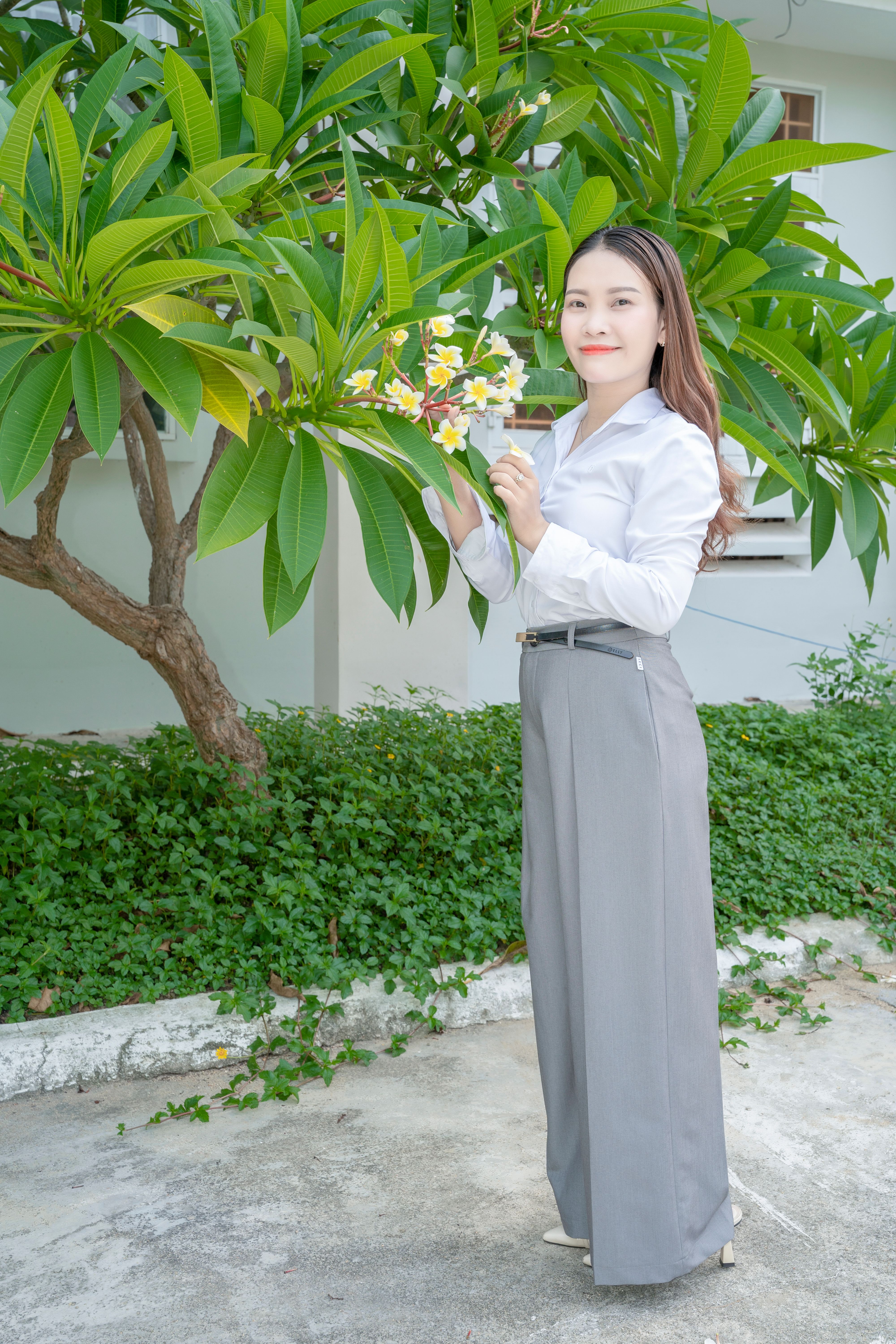 Elegant woman holding flowers, surrounded by lush green foliage, smiling warmly.