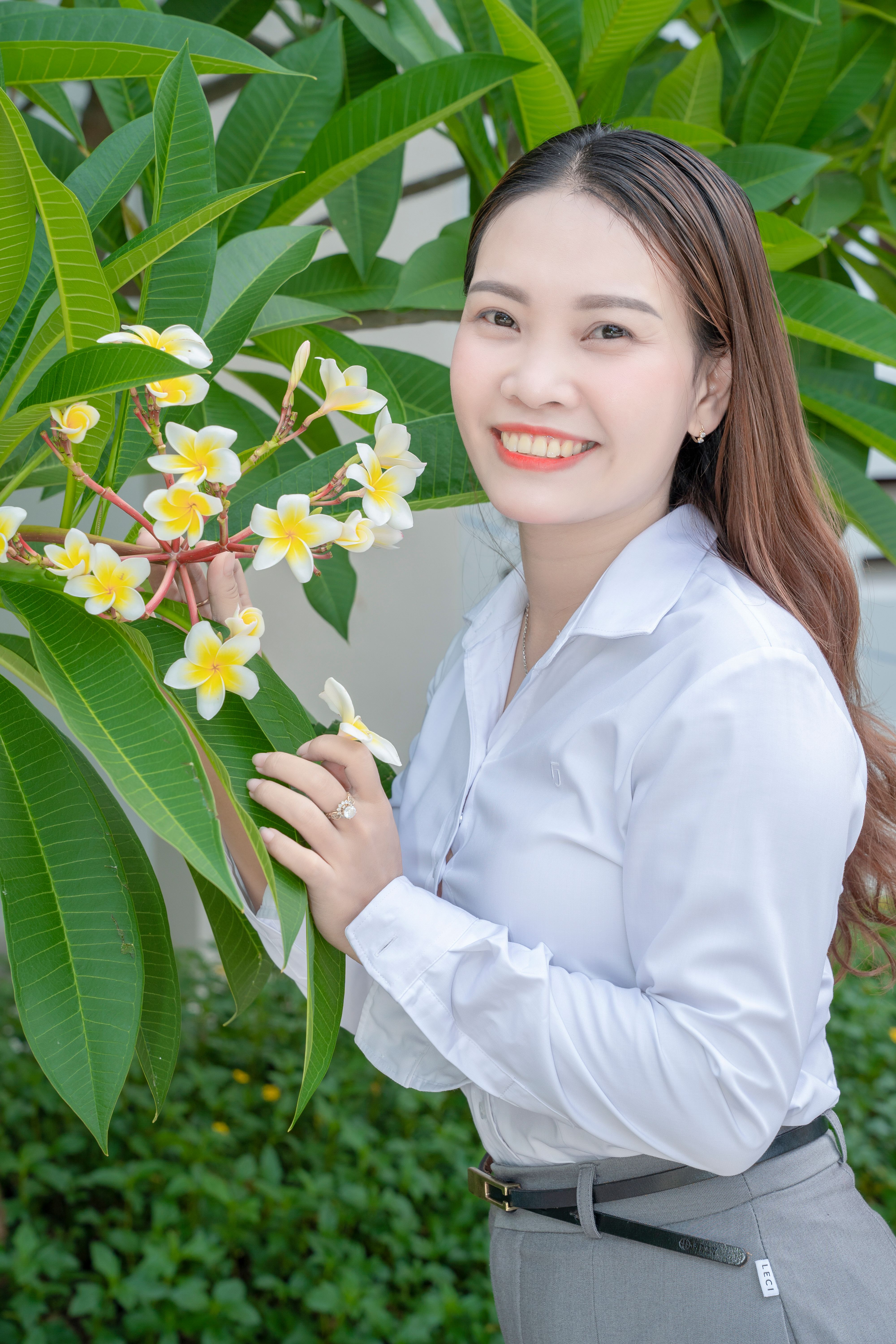 A smiling woman stands beside a frangipani tree with vibrant green leaves and white blossoms.