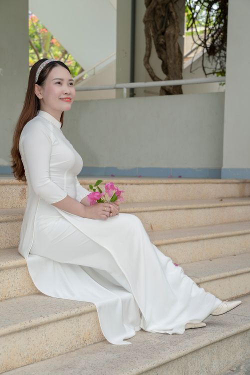 Free Side profile of a woman in a white Ao Dai holding flowers, seated on stairs. Stock Photo