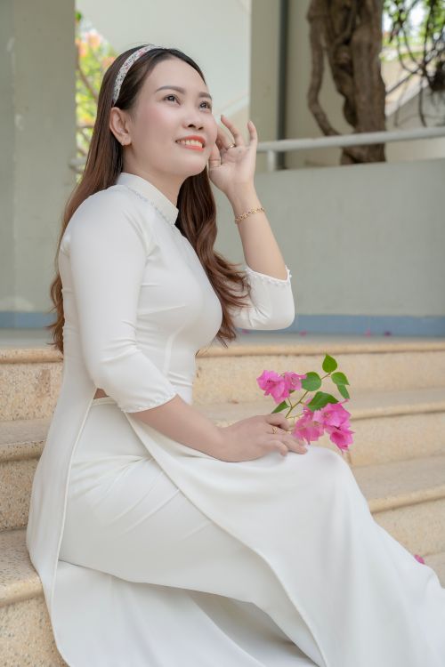 Free Serene woman in white traditional dress sitting on steps holding pink flowers. Stock Photo