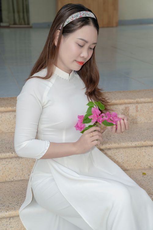 Free Serene portrait of a woman in a white Ao Dai holding pink flowers, sitting indoors. Stock Photo