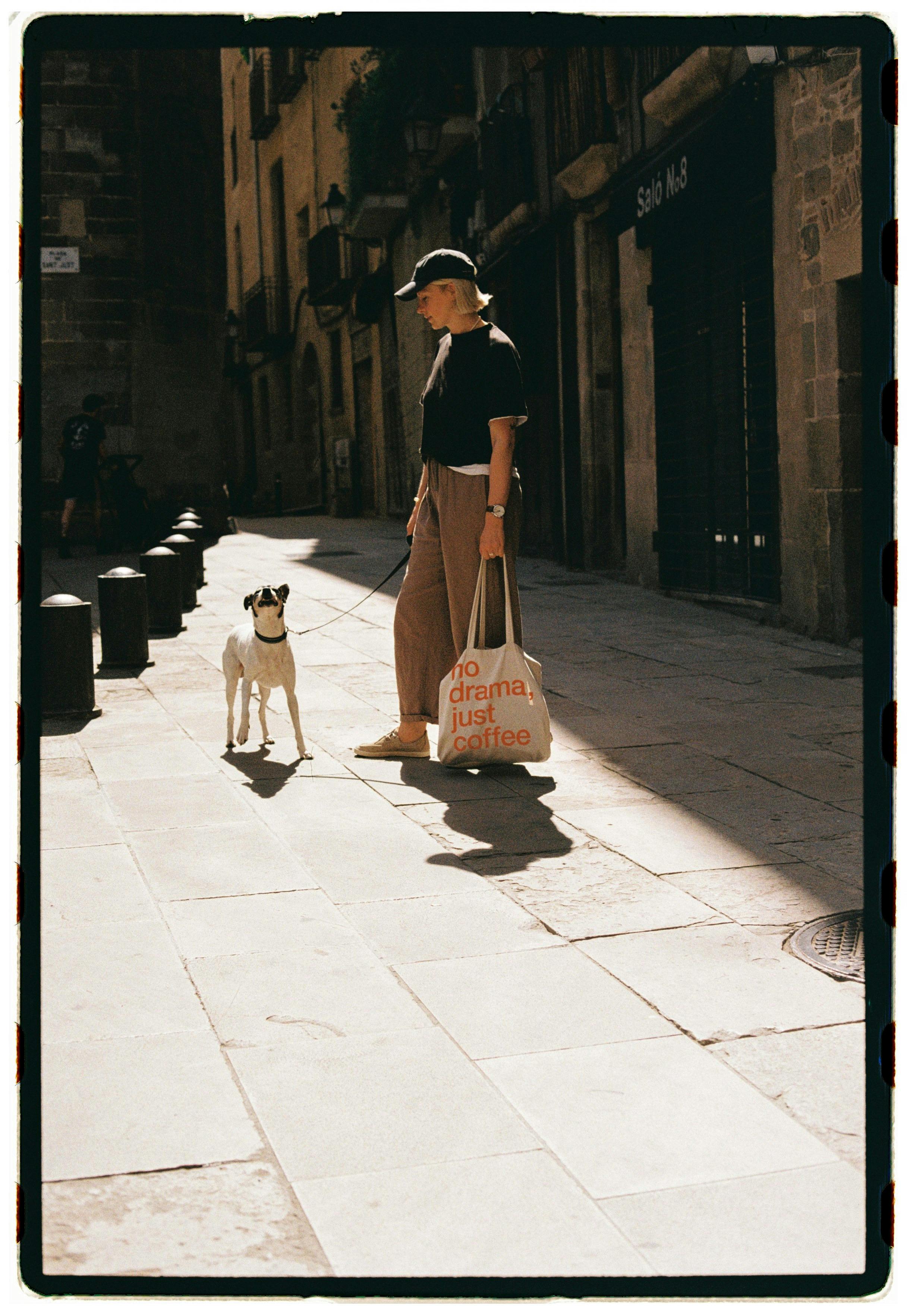 Woman and dog walking down a sunny city street, stylishly dressed holding a tote bag.