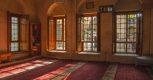 Free Warm sunlight streaming through arched windows illuminates a historic room, highlighting the rich patterns of the red carpet. Stock Photo