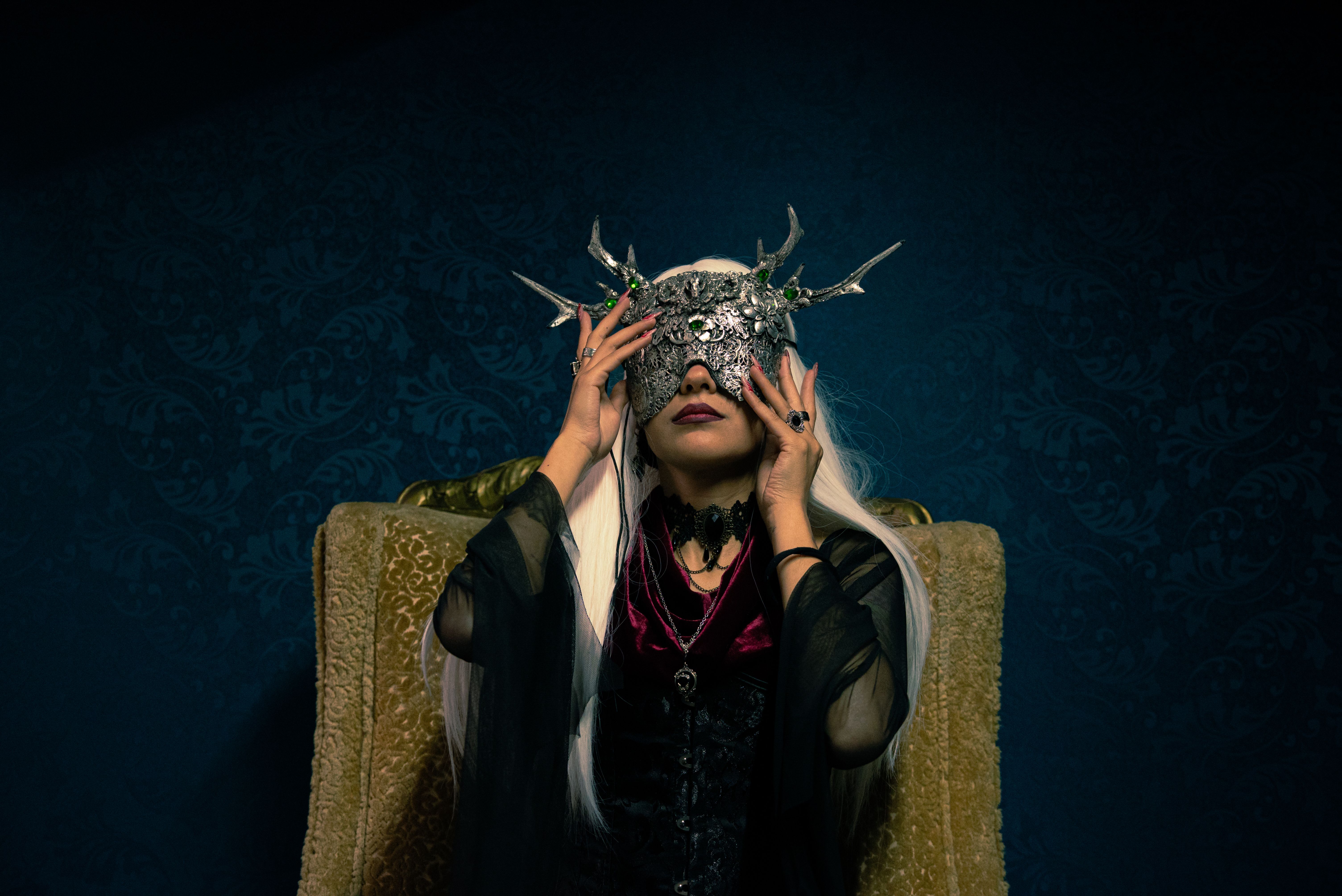 A woman in an elaborate mask with antlers, sitting on a vintage chair in a dark setting.