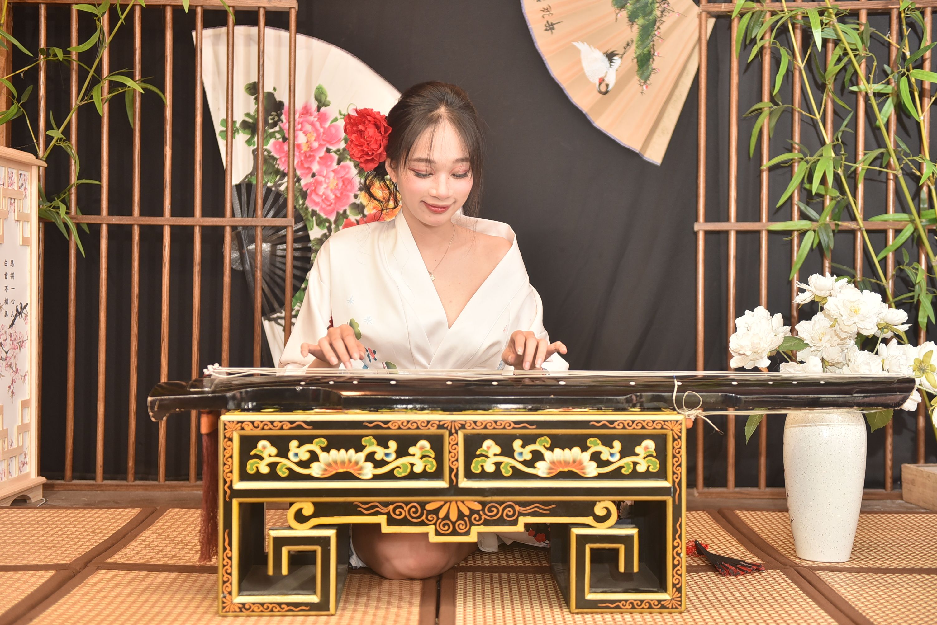 A woman in a traditional setting playing a guzheng, an ancient Chinese zither, surrounded by bamboo and floral decorations.