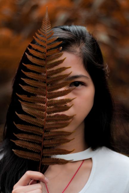 A woman holds a fern leaf in front of her face amidst autumn colors, evoking a mysterious and serene mood.