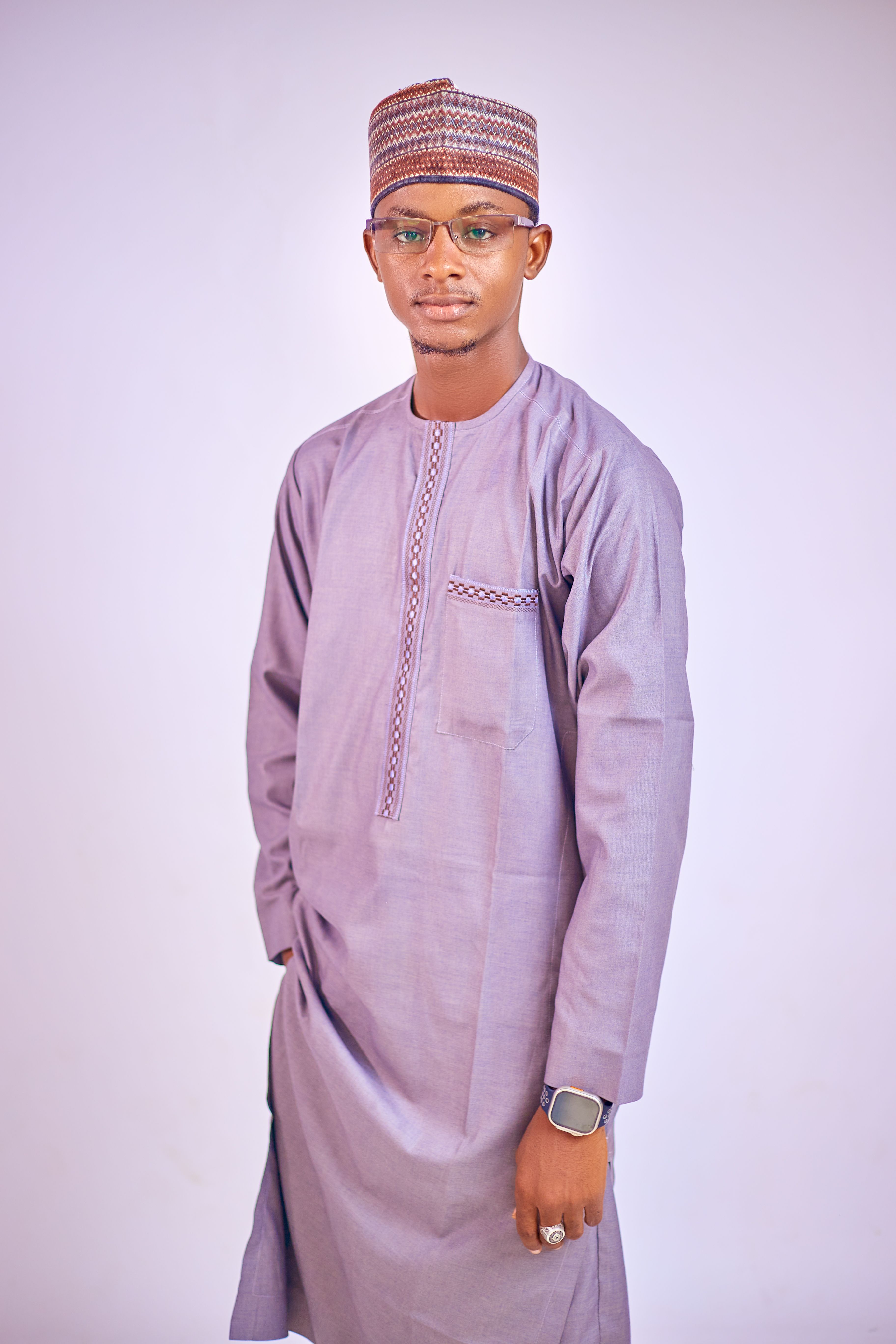 Young adult male in traditional attire with glasses and a hat on a light background.