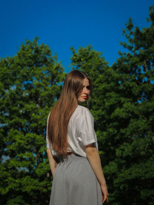 Free A young woman with long hair standing outdoors surrounded by trees on a sunny day. Stock Photo
