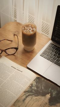 Cozy setup with iced coffee, open book, and laptop on a wooden table, ideal for work or study from home.