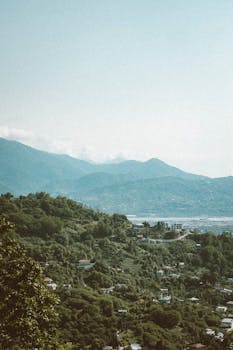 A panoramic aerial view of lush green mountains and scattered homes in Georgia, under a clear blue sky.