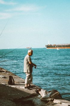 A senior man fishes from the rocky shore by a calm sea with a passing cargo ship, under a clear blue sky.