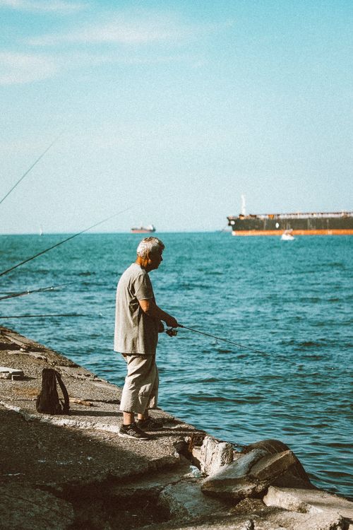 Free An elderly man fishing by the sea with ships in the distance on a sunny day. Stock Photo