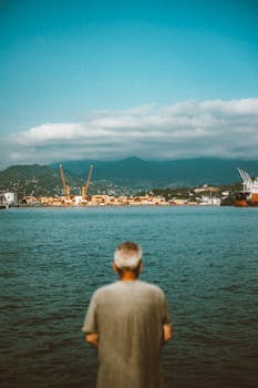 A senior man looks out over Batumi Harbor, Georgia, against a backdrop of ships and mountains.