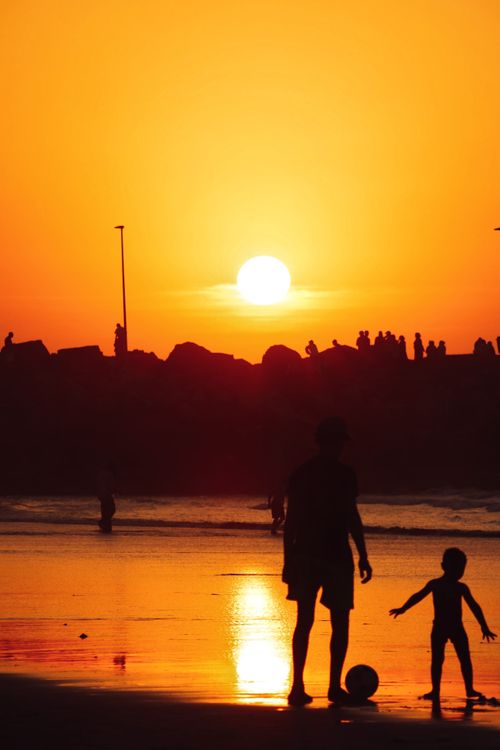 Free A beautiful sunset silhouette of a father and child playing by the beach with a soccer ball. Stock Photo