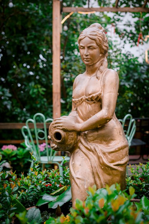 Free Golden statue of a woman with a jug in a verdant garden. Stock Photo