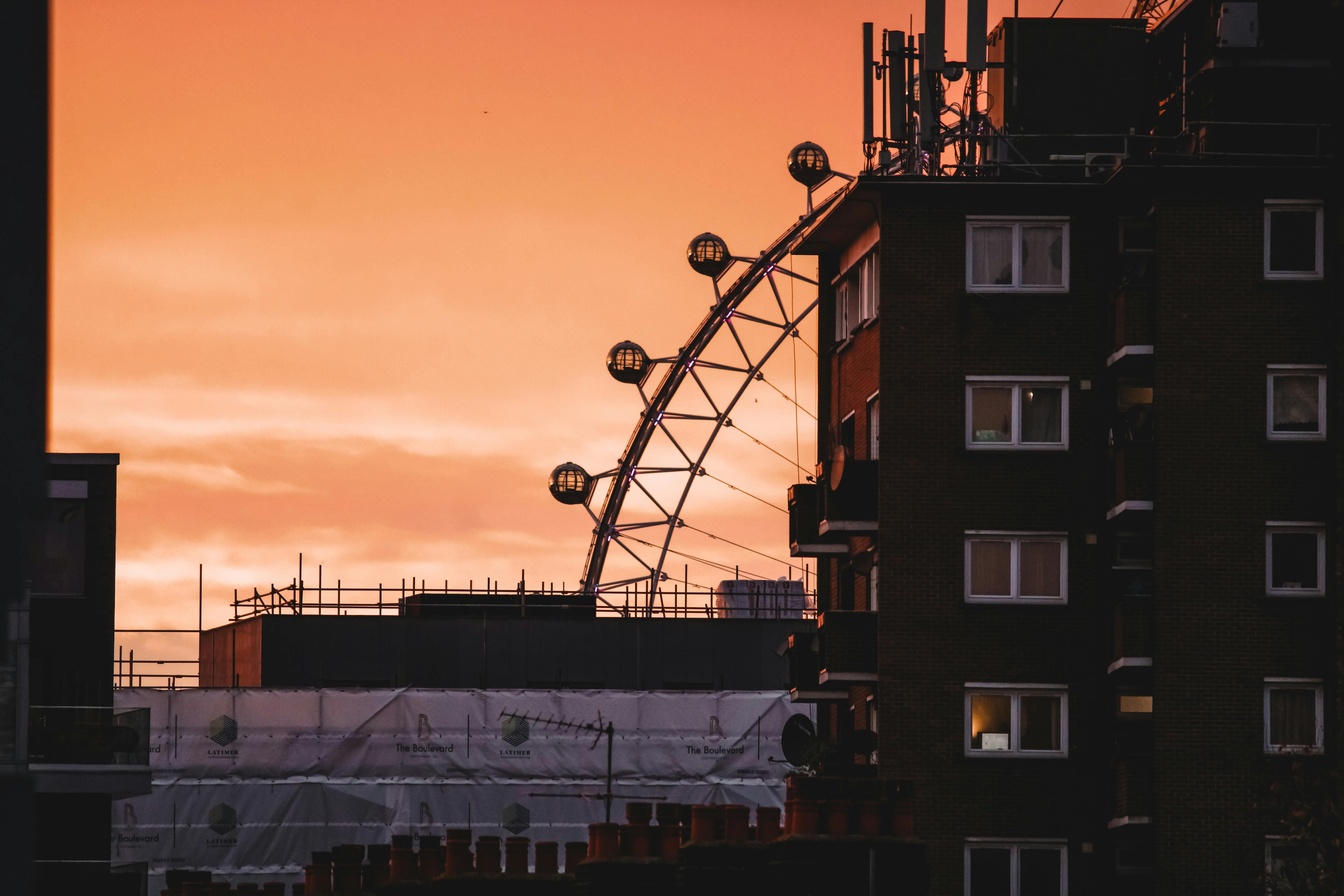 Silhouette of the London Eye against a vibrant orange sunset sky, viewed from a cityscape.