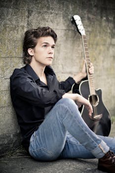 Casual portrait of a young male musician with an acoustic guitar sitting against a wall outdoors.