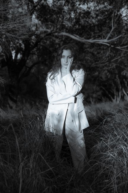A woman in a white suit poses thoughtfully in a twilight forest setting.