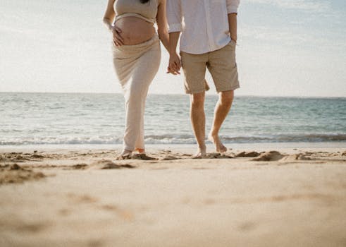 A pregnant couple holding hands while walking barefoot on a sunny beach.