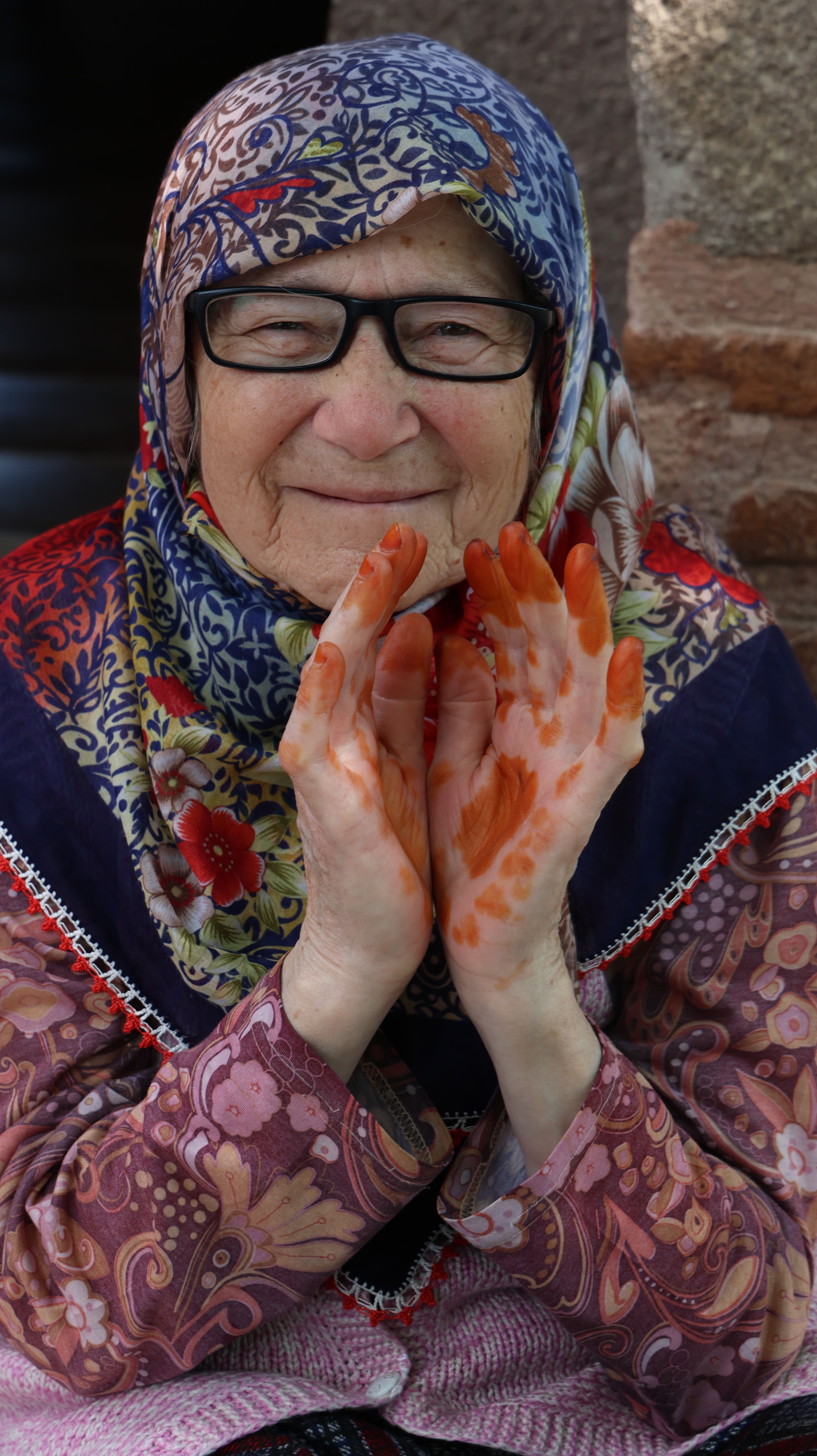 Portrait of a senior woman in colorful attire with henna-dyed hands and glasses, smiling warmly.