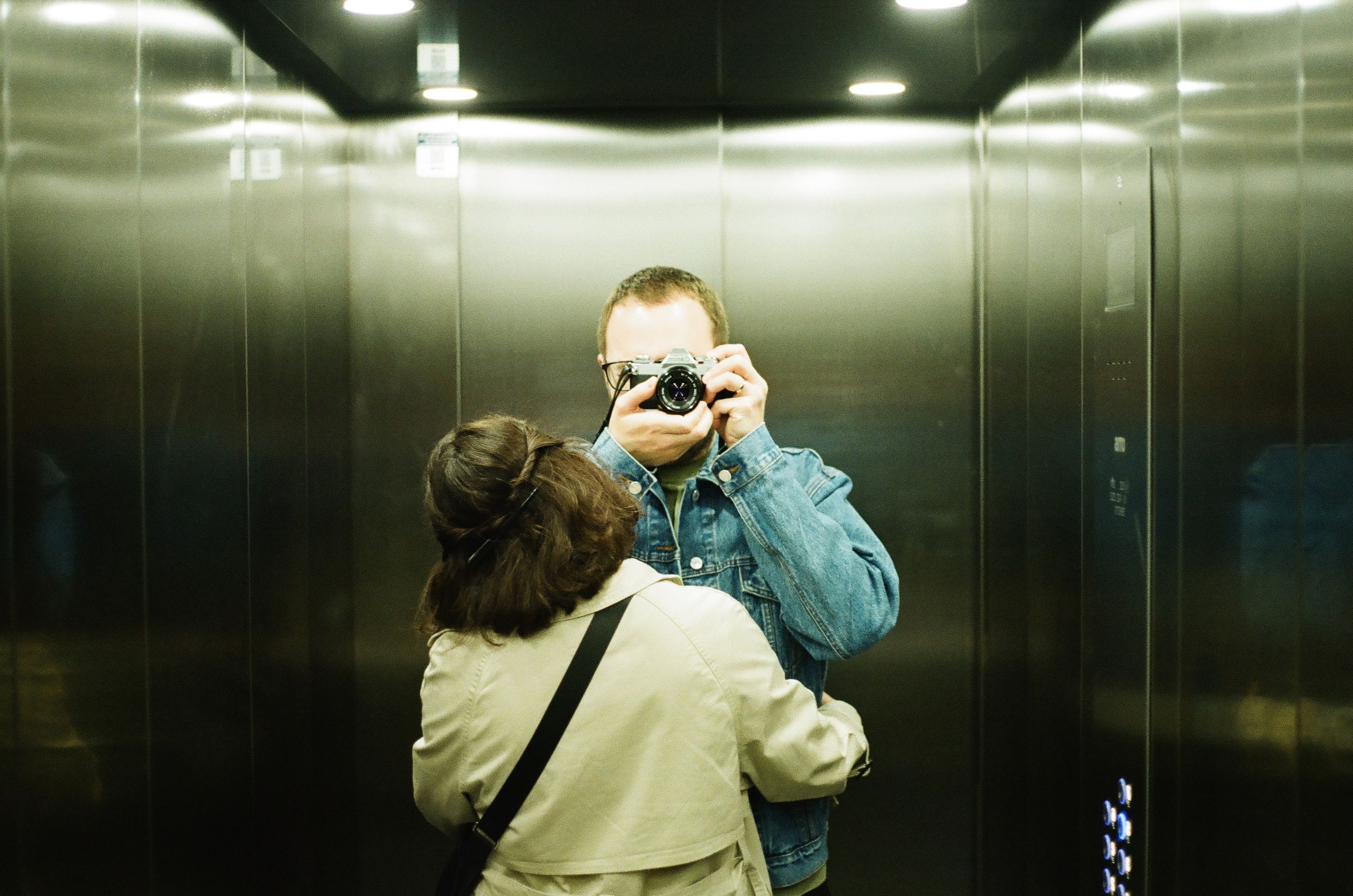 A couple captures a nostalgic mirror selfie in an elevator using a vintage film camera.