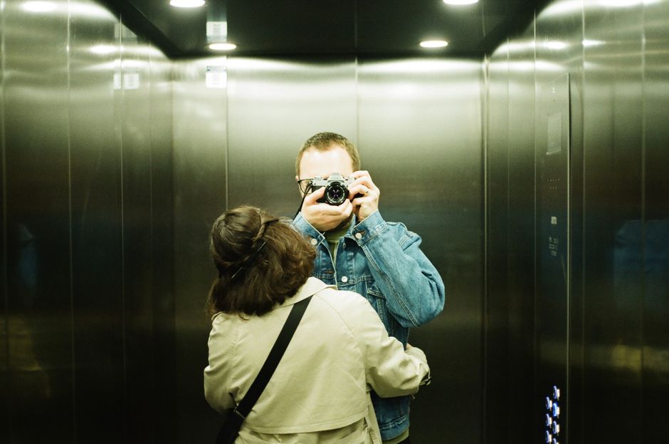 A couple captures a nostalgic mirror selfie in an elevator using a vintage film camera.