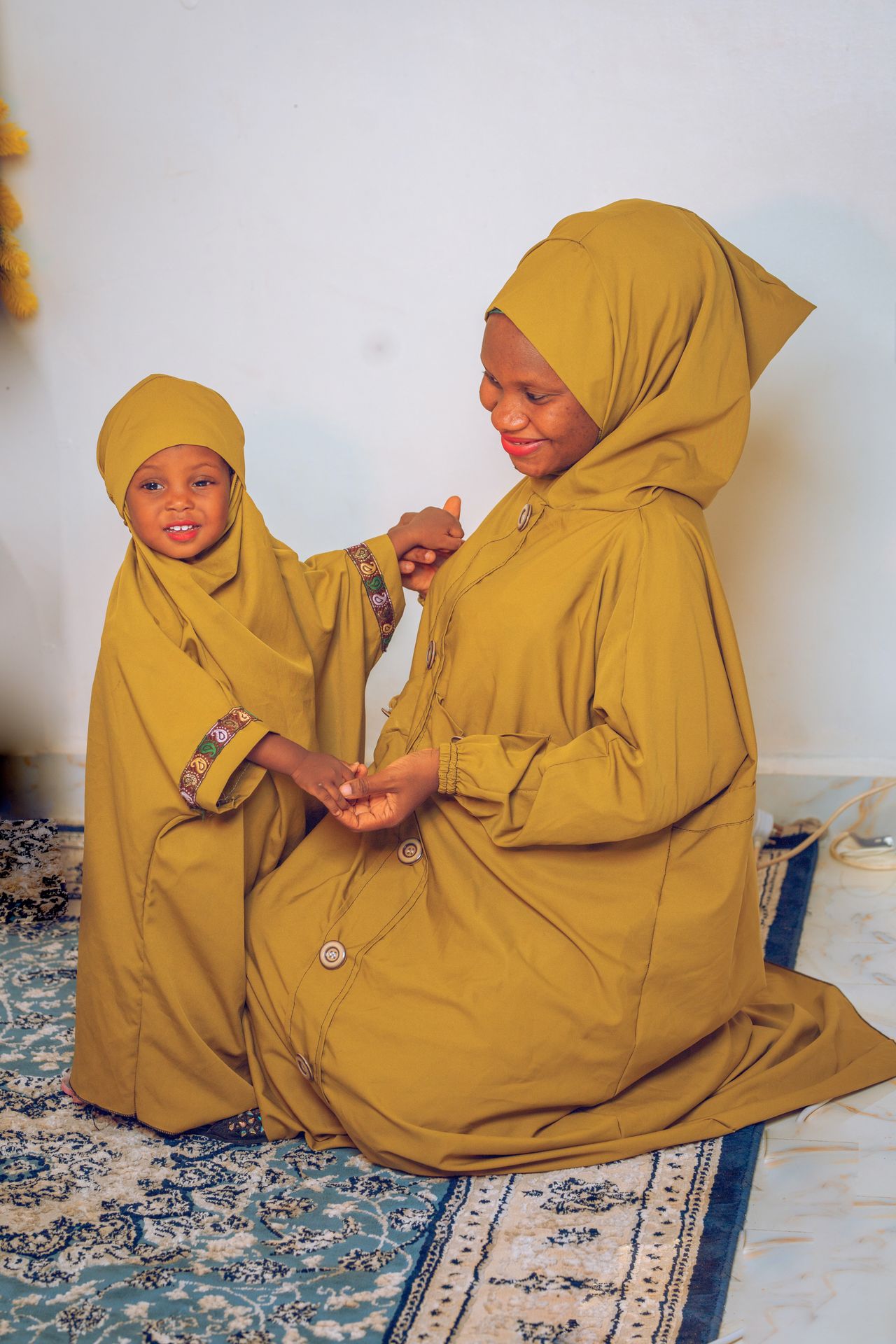 A mother and child wearing matching brown hijabs sit on a patterned rug indoors.