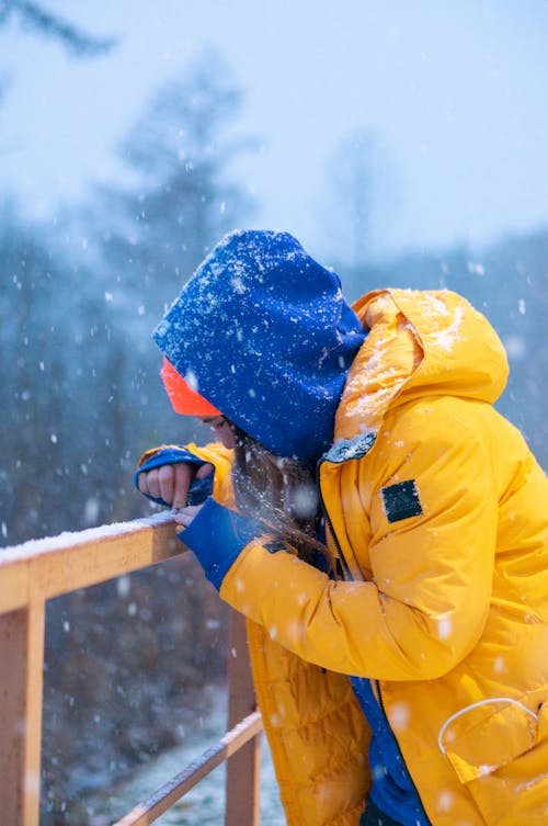 Person Wearing Yellow and Blue Jacket Leaning on Brown Fence