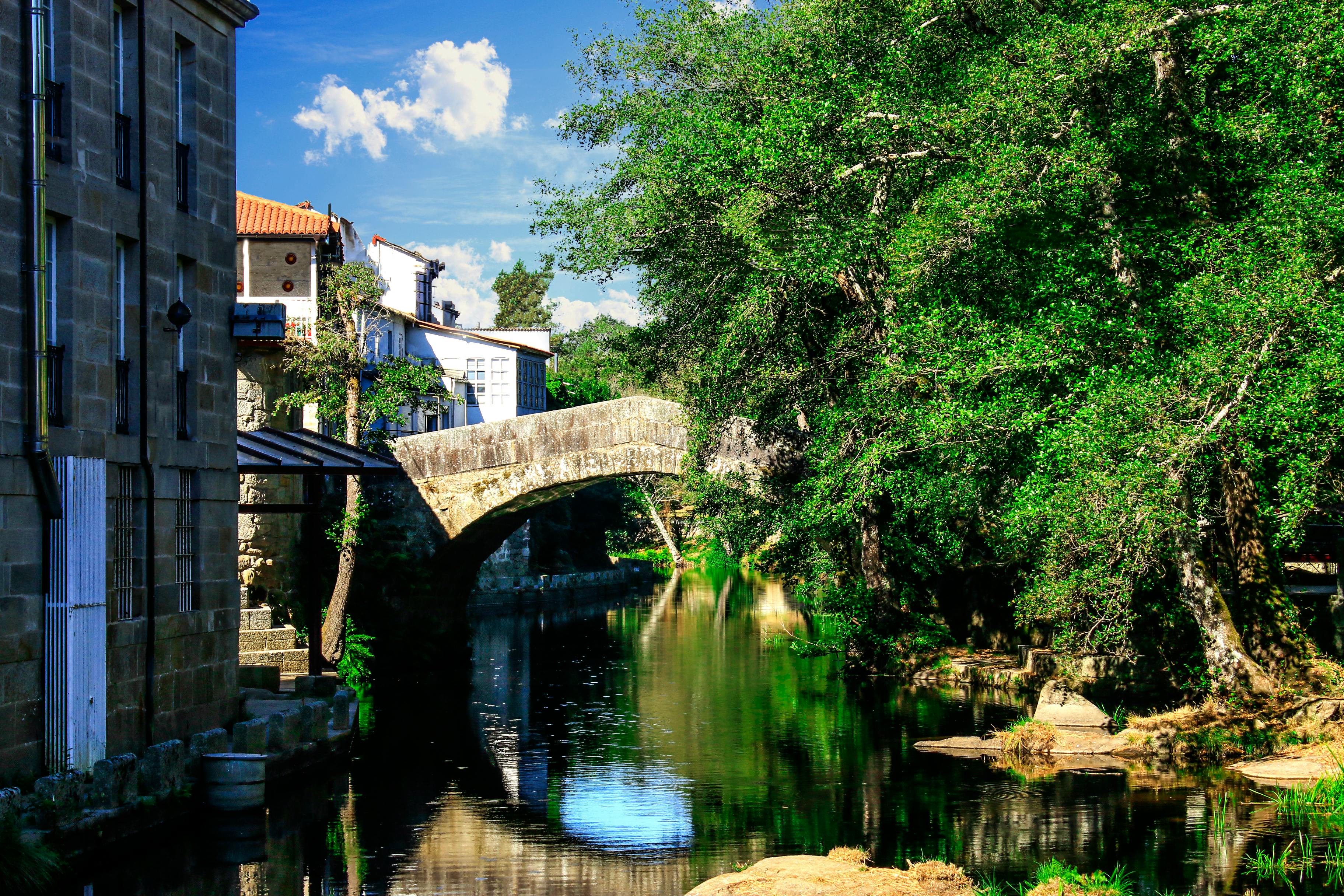 Beautiful stone bridge in Allariz, Spain, reflecting in a tranquil river surrounded by lush greenery and historic architecture.