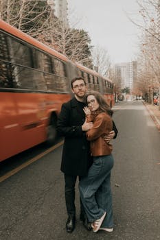 A loving couple embraces on a city street as a red bus passes by, capturing a moment of urban romance.