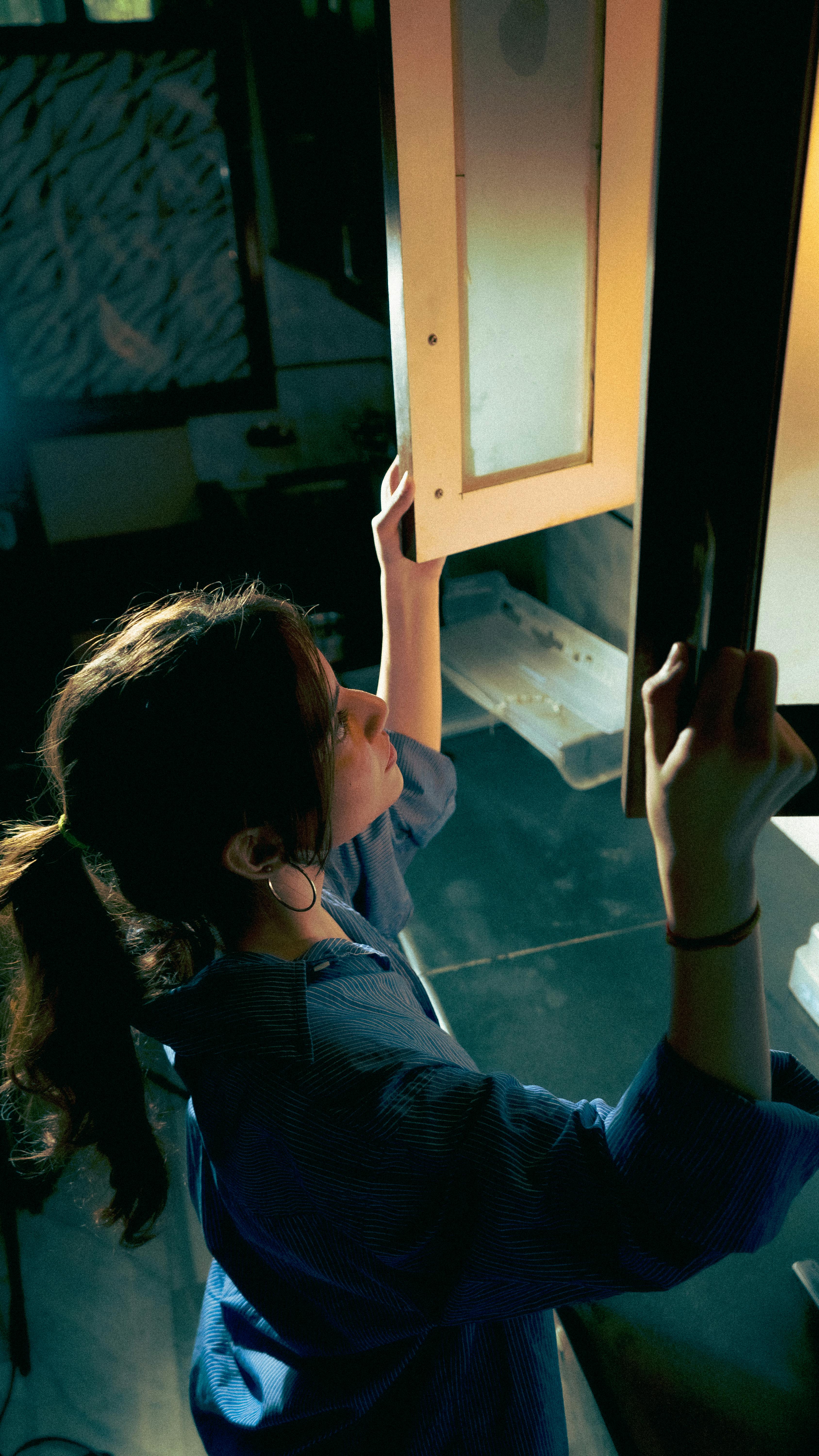 A young woman reaches for a cabinet in a moody, dimly lit kitchen, creating a dramatic scene.