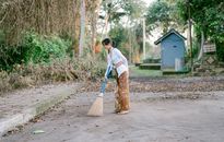 Woman cleaning a street with a broom, surrounded by trees and a serene environment.
