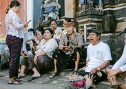 A group of Balinese adults in traditional attire relaxing and sharing outside a temple setting in daylight.