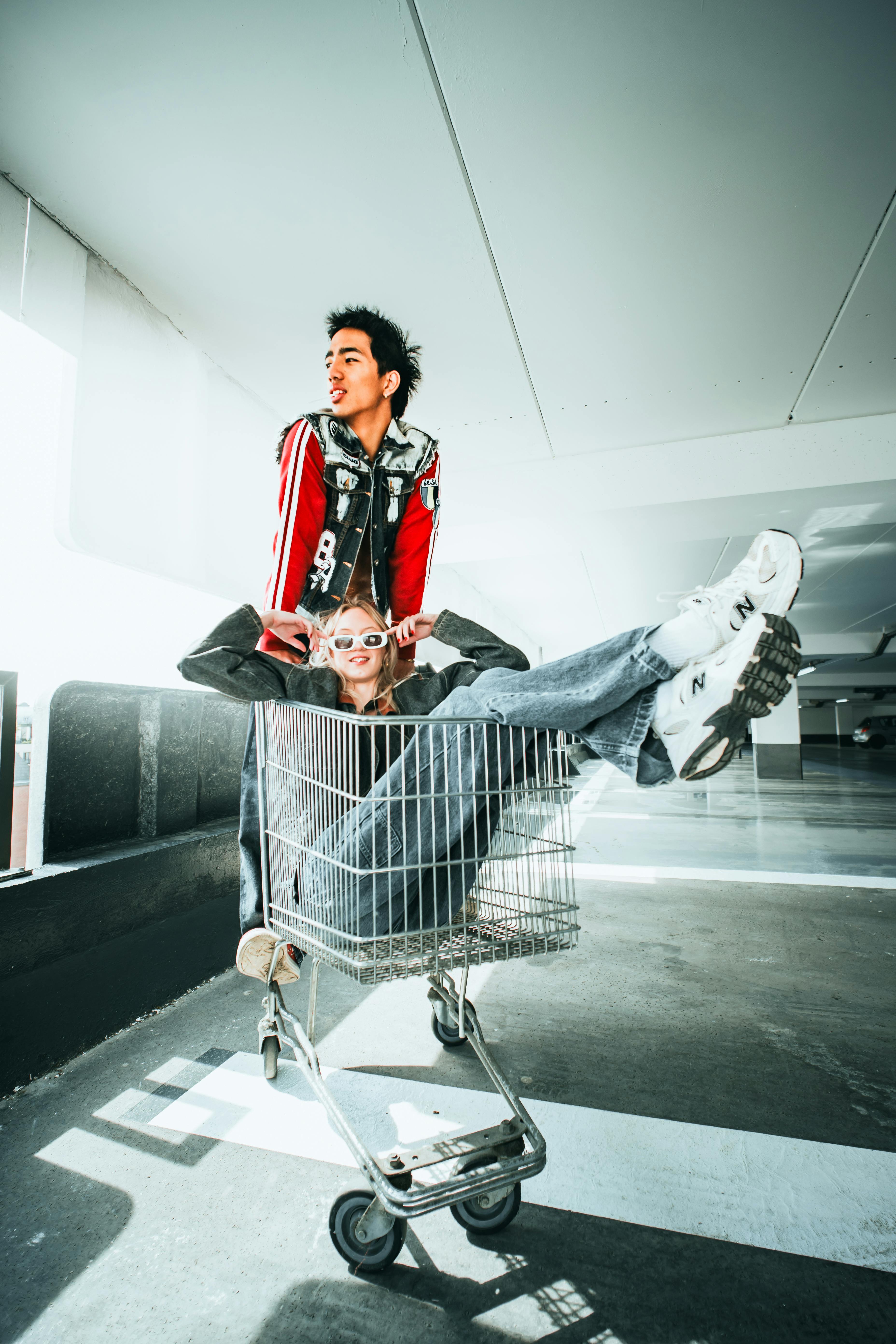 Two stylish adults enjoying a playful moment with a shopping cart in a bright urban location.