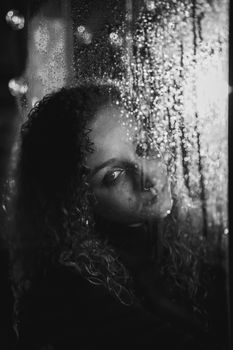 A dramatic black and white portrait of a woman looking through rain-speckled glass, evoking deep emotions.