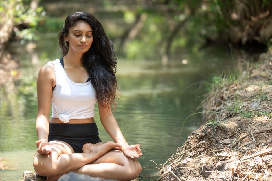 A serene woman meditating beside a peaceful river surrounded by nature